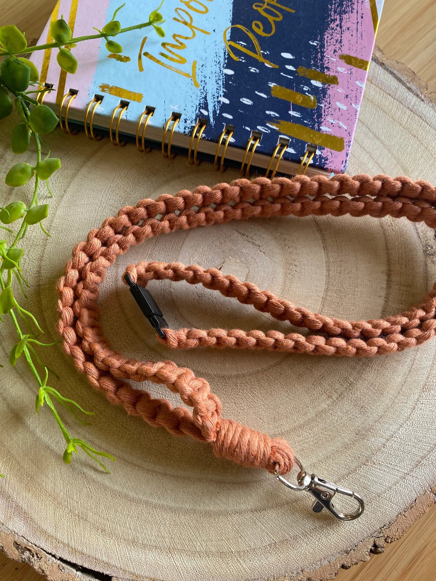 A braided, rust-colored Macra-Made-With-Love Eco friendly lanyard badge holder - 34” with a metal clasp is coiled on a wooden surface. Beside it lies a vibrant notebook with a spiral binding, partially covered by trailing green plant leaves.