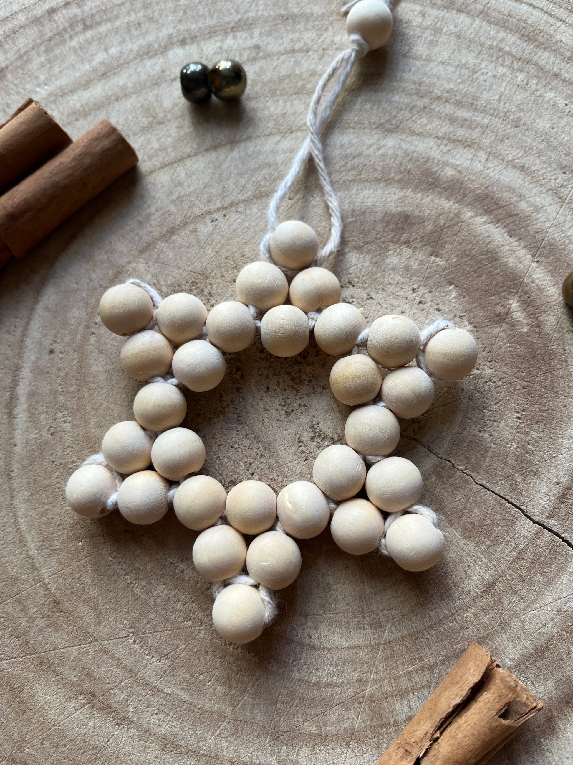 Close up of a star-shaped decorative item made of small wooden beads and beige yarn, on a wooden surface with cinnamon sticks and gold beads for decoration.