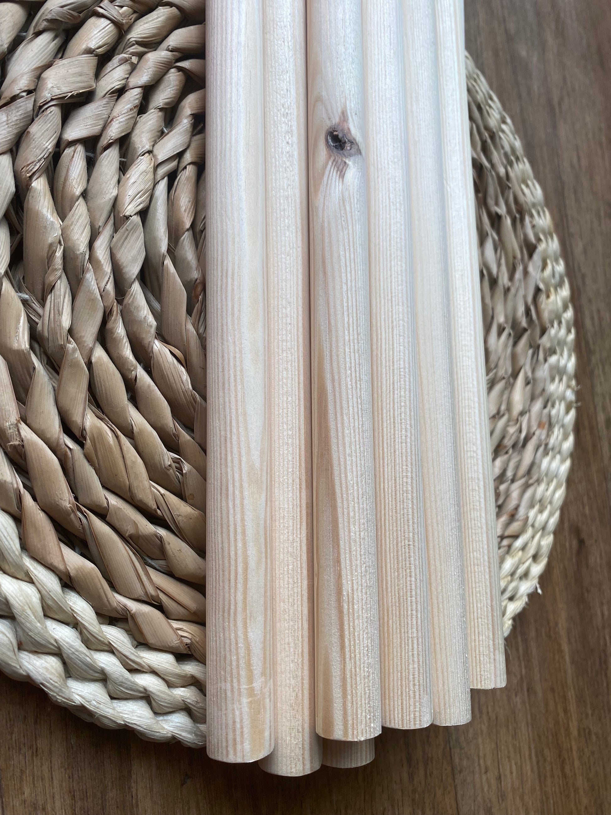 Close up of a pile of natural pine wooden dowels for macramé and crafts laid across a round wicker mat. 
