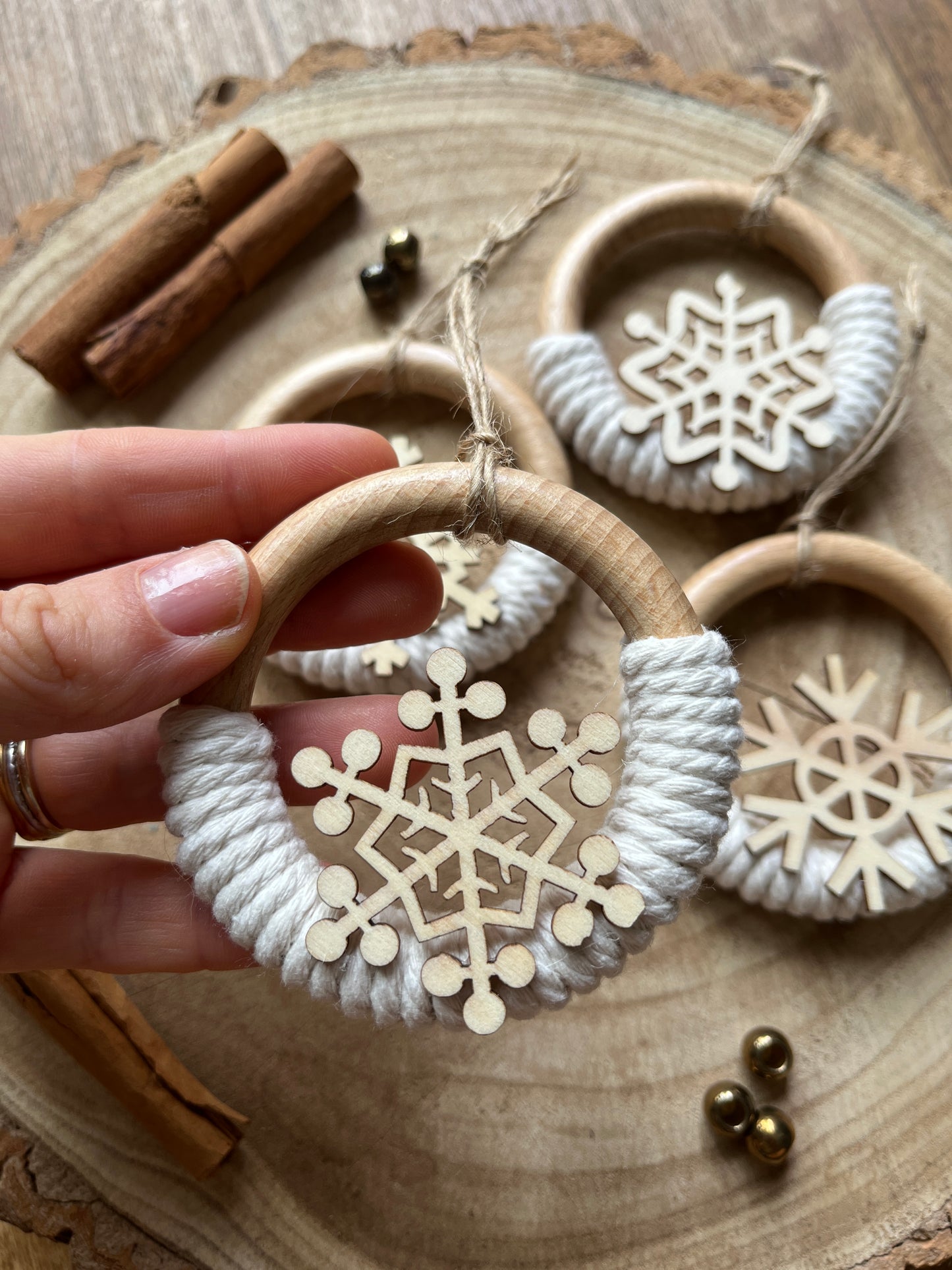 A hand holding a decorative wooden ornament, made on a wooden ring with natural yarn and finished with a wooden snowflake design, there are three other ornaments displayed on a rustic wooden surface in the background. 