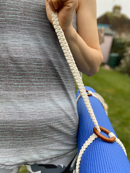 A woman stands outside in exercise clothes, with a blue yoga mat held with a macrame yoga mat strap on her shoulder. 