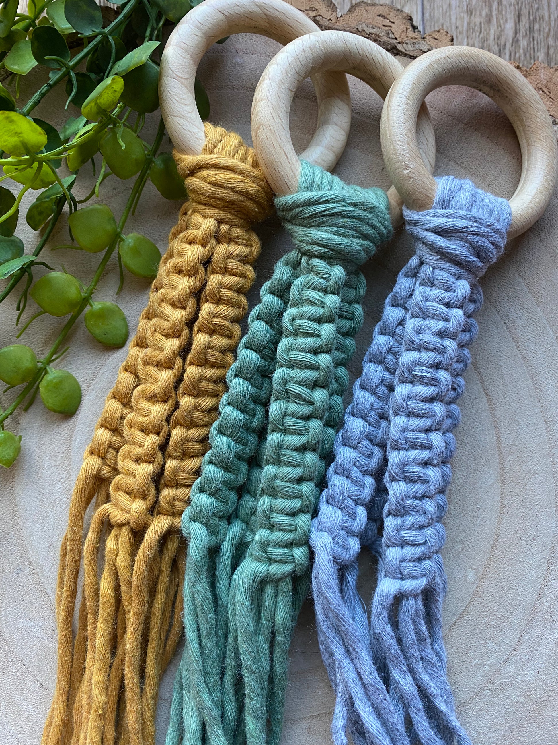Three macrame plant hangers, made with mustard, green and grey cotton yarn on light coloured wooden rings, lay next to each other on a wooden surface next to some green plant vines. 