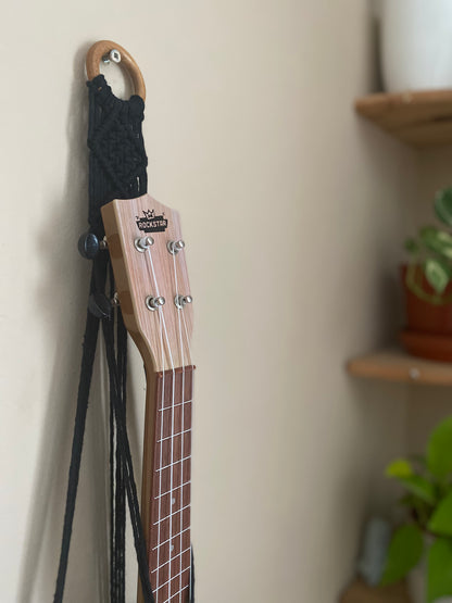 A light brown ukulele sits inside of a handmade macrame wall hanging ukulele hanger, which is made with black recycled cotton, and a repurposed wooden hanging ring. 