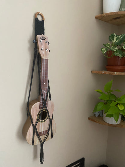 A light brown ukulele sits inside of a handmade macrame wall hanging ukulele hanger, which is made with black recycled cotton, and a repurposed wooden hanging ring. Two green leafy plants sit on shelves beside the hanger.