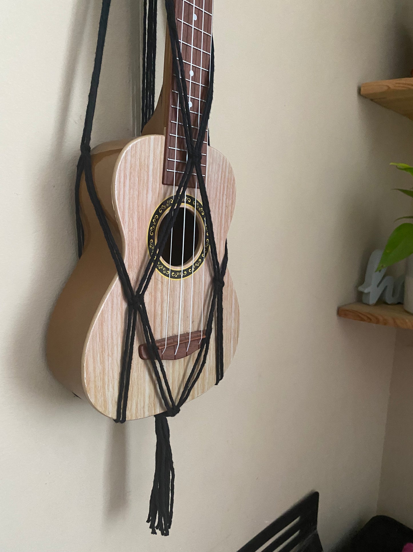 A light brown ukulele sits inside of a handmade macrame wall hanging ukulele hanger, which is made with black recycled cotton. The photo focuses on the bottom part of the ukulele in the hanger. 