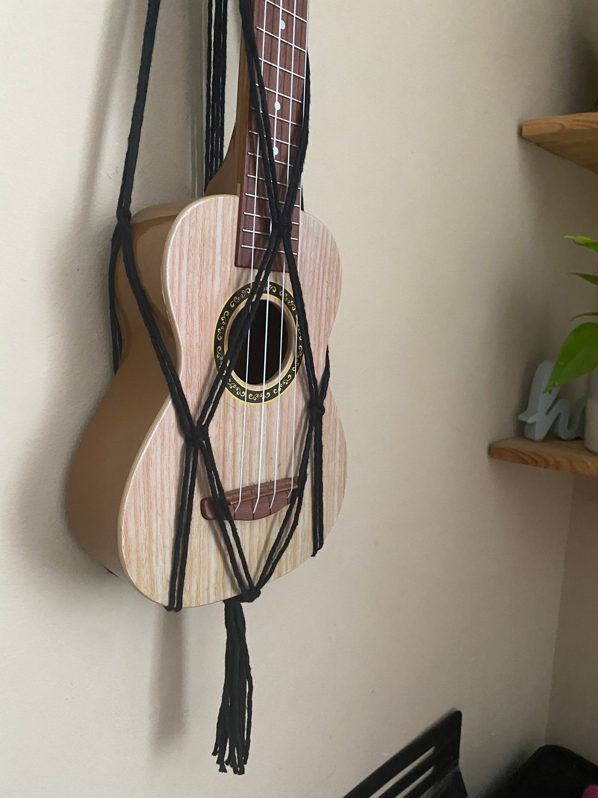 A light brown ukulele sits inside of a handmade macrame wall hanging ukulele hanger, which is made with black recycled cotton. The photo focuses on the bottom part of the ukulele in the hanger. 