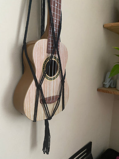 A light brown ukulele sits inside of a handmade macrame wall hanging ukulele hanger, which is made with black recycled cotton. The photo focuses on the bottom part of the ukulele in the hanger. 