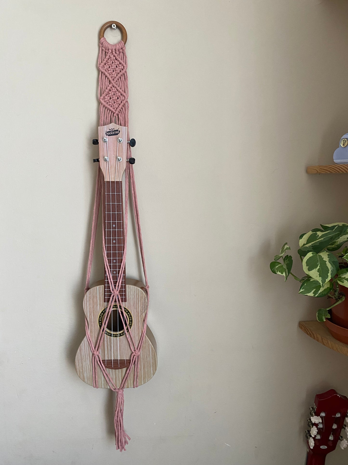A light brown ukulele sits inside of a handmade macrame wall hanging ukulele hanger, which is made with blush pink recycled cotton, and a repurposed wooden hanging ring. 