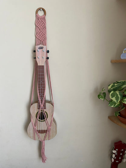 A light brown ukulele sits inside of a handmade macrame wall hanging ukulele hanger, which is made with blush pink recycled cotton, and a repurposed wooden hanging ring. 