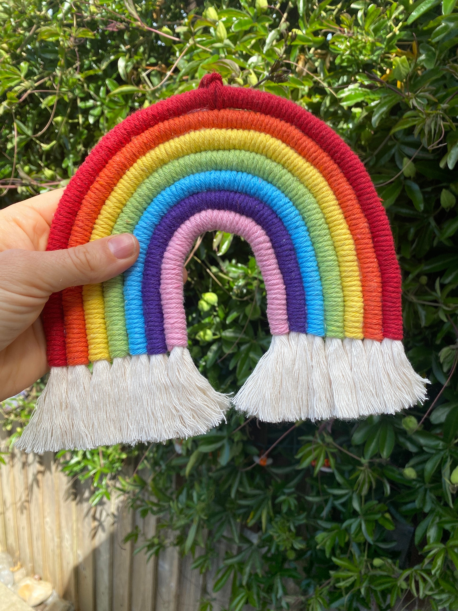 A hand holding a macrame rainbow made with 7 arches of colourful yarn, with white tassels at the bottom outside with lush green plants in the background. 