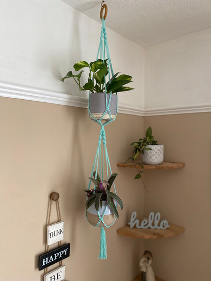 Two trailing plants in ceramic pots, suspended vertically in a large double macramé plant hanger, made with mint coloured yarn and a wooden hanging ring, in the corner of a room with beige walls. There is a set of corner shelves in the background.