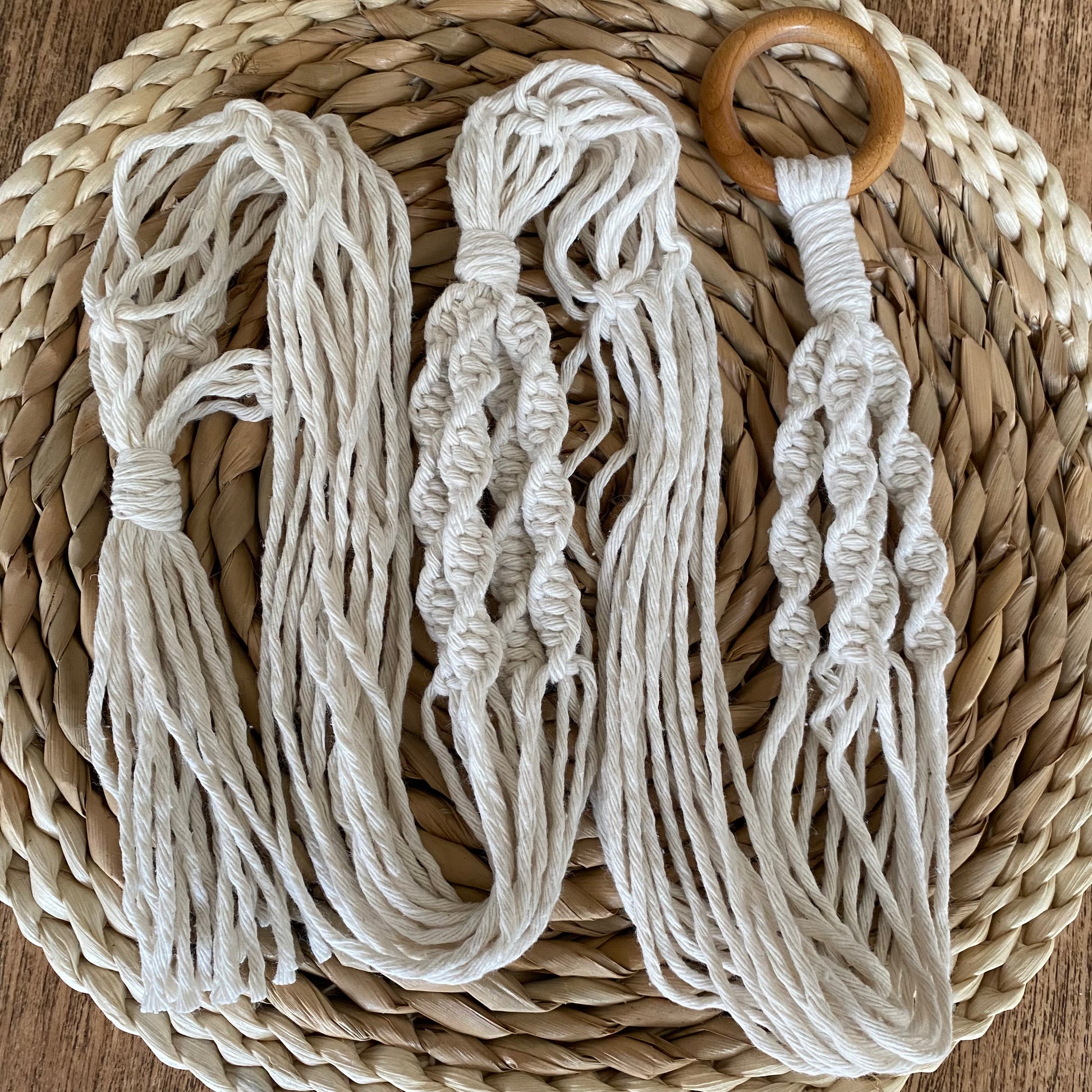 A macrame plant hanger made with natural yarn and a wooden ring, is coiled on a round wicker placemat. 