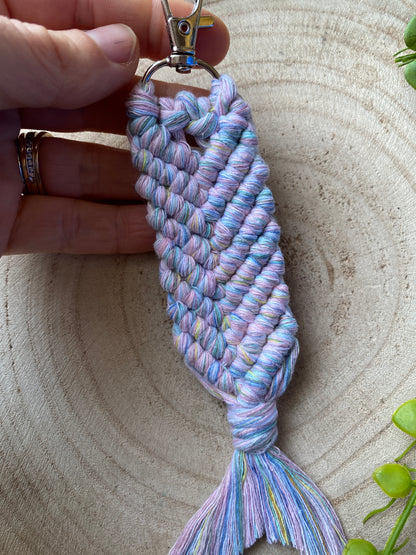 A hand holding a Macra-Made With Love handmade macrame keyring, in the shape of a mermaid tail, made with soft pastel coloured recycled cotton, finished with a metal lobster clasp. 