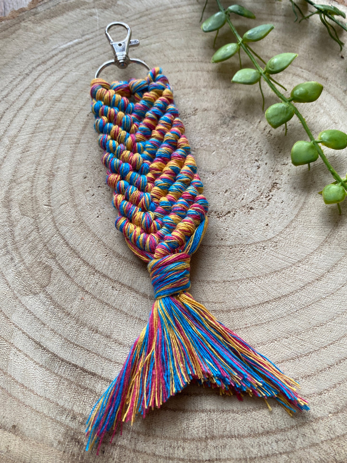 A handmade macrame keyring, knotted using brightly coloured recycled cotton in the shape of a mermaids tail, and finished with a metal lobster clasp. Displayed on a wooden surface with some green plant vines. 
