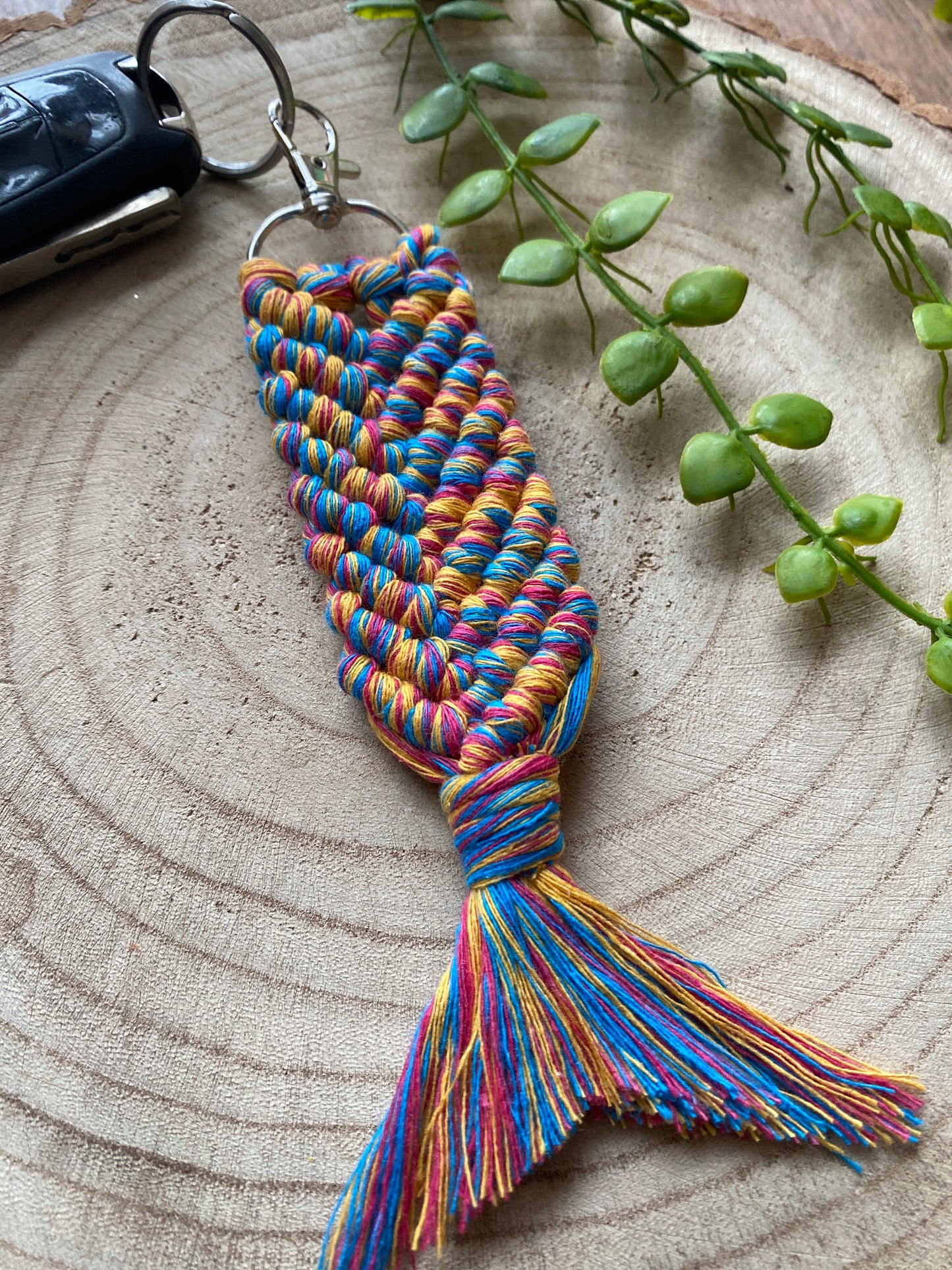 A handmade macrame keyring, knotted using brightly coloured recycled cotton in the shape of a mermaids tail, and finished with a metal lobster clasp. Shown attached to a single black car key, on a wooden surface with some green plant vines nearby.