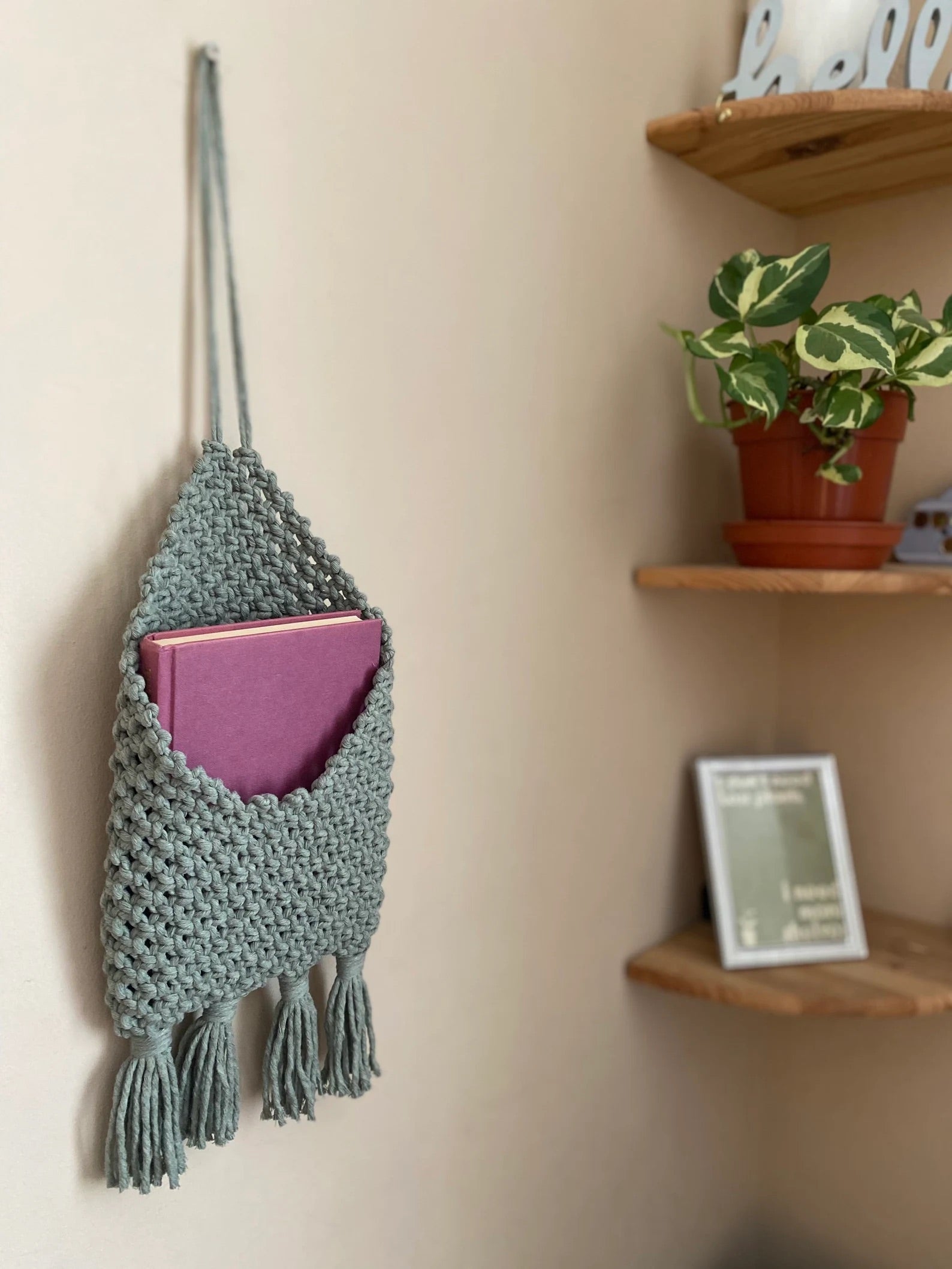 A handmade Macra-Made-With-Love Bedside storage pouch book sleeve in light blue holds a closed pink book. Featuring a textured pattern with tassels at the bottom, it adds charm to any space. In the background, an eco-friendly bedroom storage setup includes a wooden shelf with a potted plant and a small picture frame.