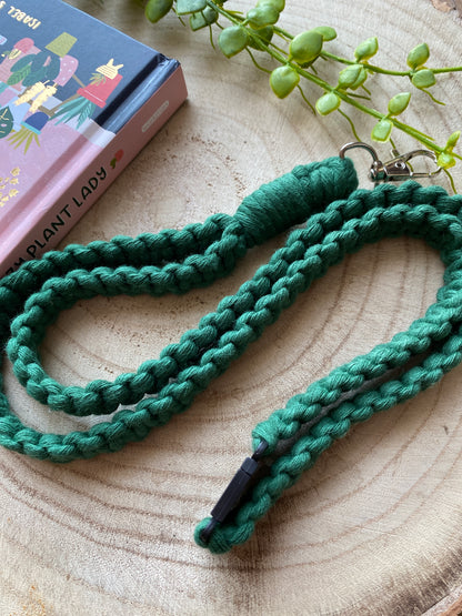 A green, handmade lanyard with a metal clip and black plastic clasp is laid out on a wooden surface. A book titled "Plant Lady" with illustrations of plants and a partial view of a succulent plant are visible in the background, perfect for holding your Macra-Made-With-Love Eco friendly lanyard badge holder - 34”.