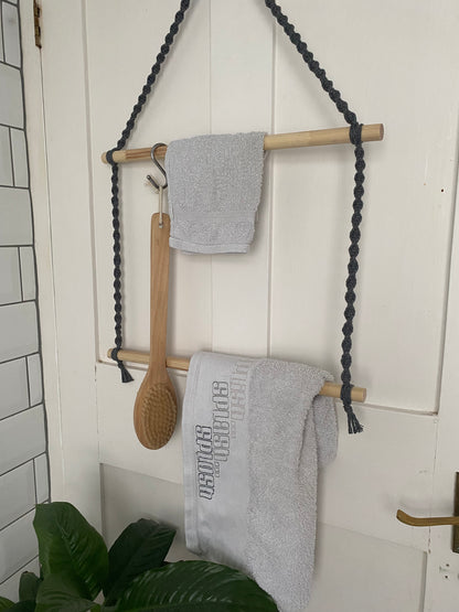 A Two tier bathroom towel holder by Macra-Made-With-Love with two horizontal rods, suspended by black braided ropes, hangs on a white door. A small gray towel and a wooden back brush hang on the top rod, while a larger gray towel with text hangs on the bottom rod. This eco-friendly towel rail adds a touch of nature to your bathroom decor. A plant is visible at the bottom.