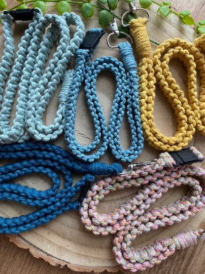 A variety of colorful, braided dog leashes and Macra-Made-With-Love Eco friendly lanyard badge holders - 34” are neatly arranged on a wooden surface. The leashes come in shades of light blue, dark blue, teal, yellow, and a multicolored pastel. Each leash has a metal clasp for attaching to a collar. Green leaves are visible in the background.