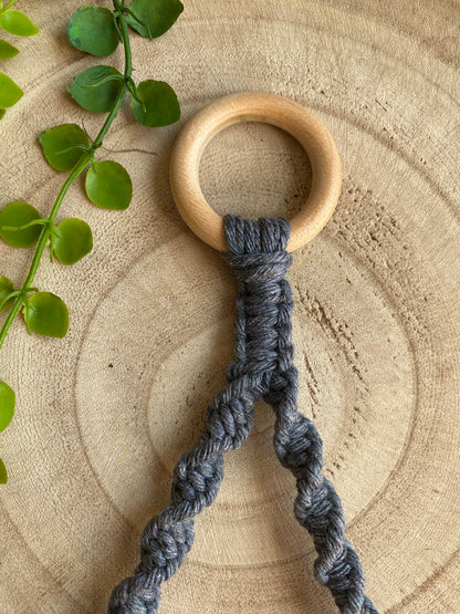 A Single toilet roll holder made with gray cord from Macra-Made-With-Love is seen against a wooden background. The eco-friendly decor piece features a wooden ring at the top and spiral knots along its length. A small green plant is partially visible to the left.