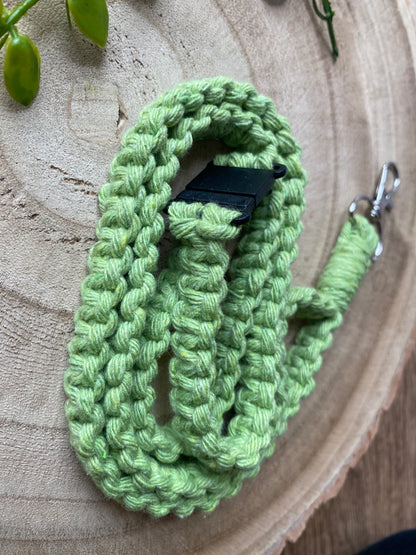 A tightly coiled, light green crocheted **Eco friendly lanyard badge holder - 34”** from **Macra-Made-With-Love**, with a silver clasp and a black plastic clip, reminiscent of an eco-friendly lanyard, rests on a wooden surface with a couple of small succulent leaves partly visible in the background.