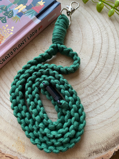 A green, woven rope dog leash with a metal clasp is coiled neatly on a wooden surface. The leash features a black plastic fastener in the middle, resembling the quality of an Eco friendly lanyard badge holder - 34” from Macra-Made-With-Love. In the upper left corner, a book titled "Crazy Plant Lady" with a colorful cover is partially visible.