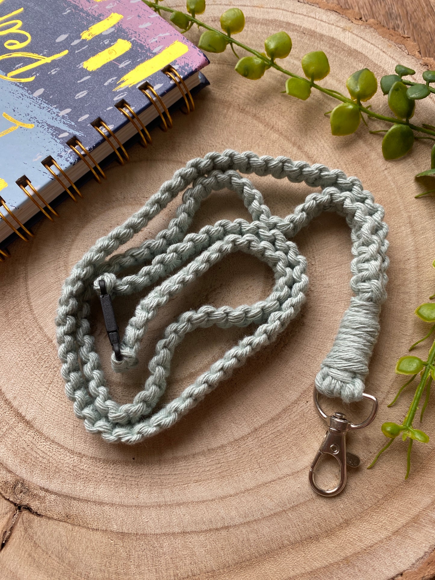 A handwoven, light gray, eco-friendly lanyard badge holder - 34” from Macra-Made-With-Love with a metal clasp lies coiled on a wooden surface. To the top left is a colorful spiral-bound notebook with abstract designs. Green leafy stems are scattered around the lanyard and notebook, adding a natural touch to the scene.