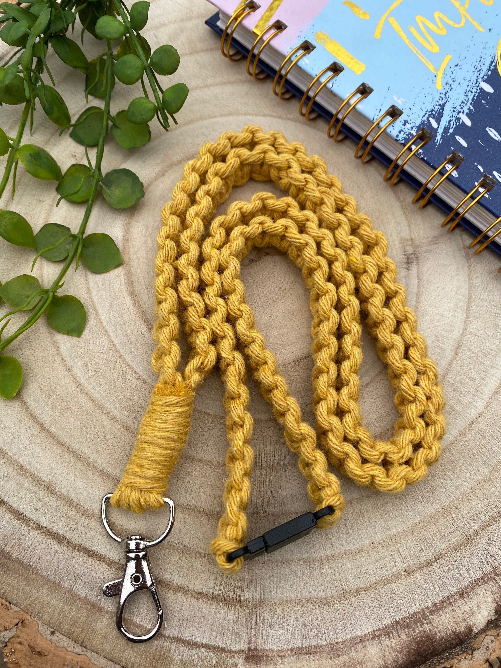 A handmade yellow braided lanyard with a metal clip lies coiled on a wooden surface. Beside it is a spiral-bound notebook with a colorful cover, and partially visible green leaves add a touch of nature to the scene. This Eco friendly lanyard badge holder - 34” from Macra-Made-With-Love makes for both a stylish accessory and an ID badge holder.