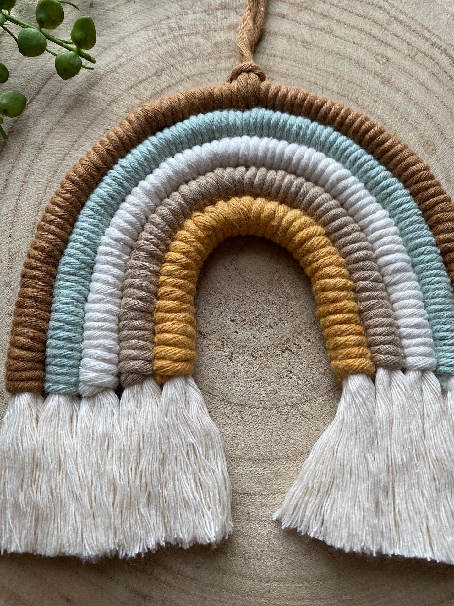 A handmade, Neutral Rainbow from Macra-Made-With-Love with earthy tones of brown, light blue, light grey, mustard yellow, and white. Adorned with white tassels hanging from the ends and laid on a wooden surface with a sprig of green leaves in the corner, it's perfect as a baby shower gift crafted using sustainable practices.