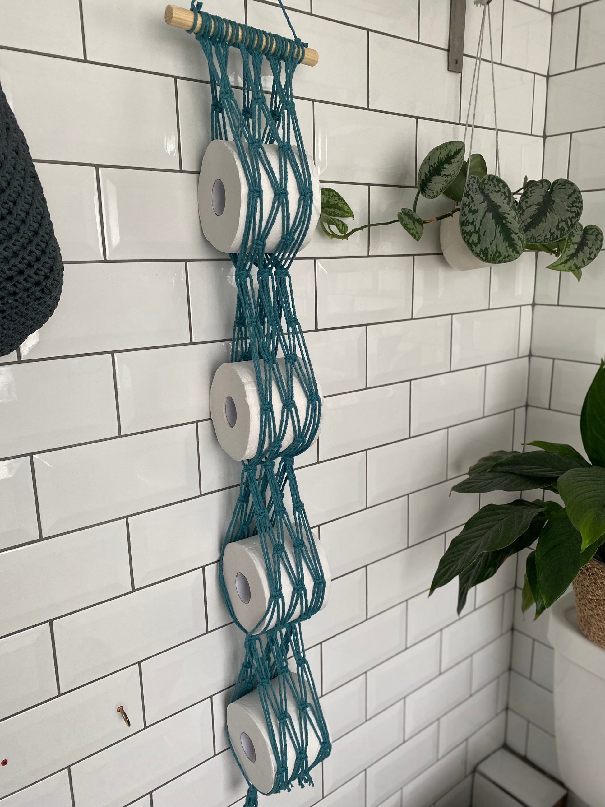 A bathroom wall with a white subway tile backdrop features a teal Macra-Made-With-Love Toilet paper wall mounted storage containing five rolls of toilet paper. Nearby, green potted plants add a touch of nature to the bathroom decor, creating an eco-friendly ambiance.