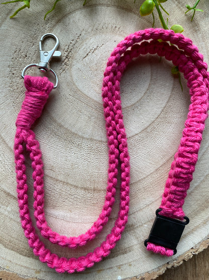 A bright pink braided, Macra-Made-With-Love Eco friendly lanyard badge holder - 34” with a silver metal clip on one end and a black plastic clasp on the other, placed on a wooden surface with green plant accents in the background.