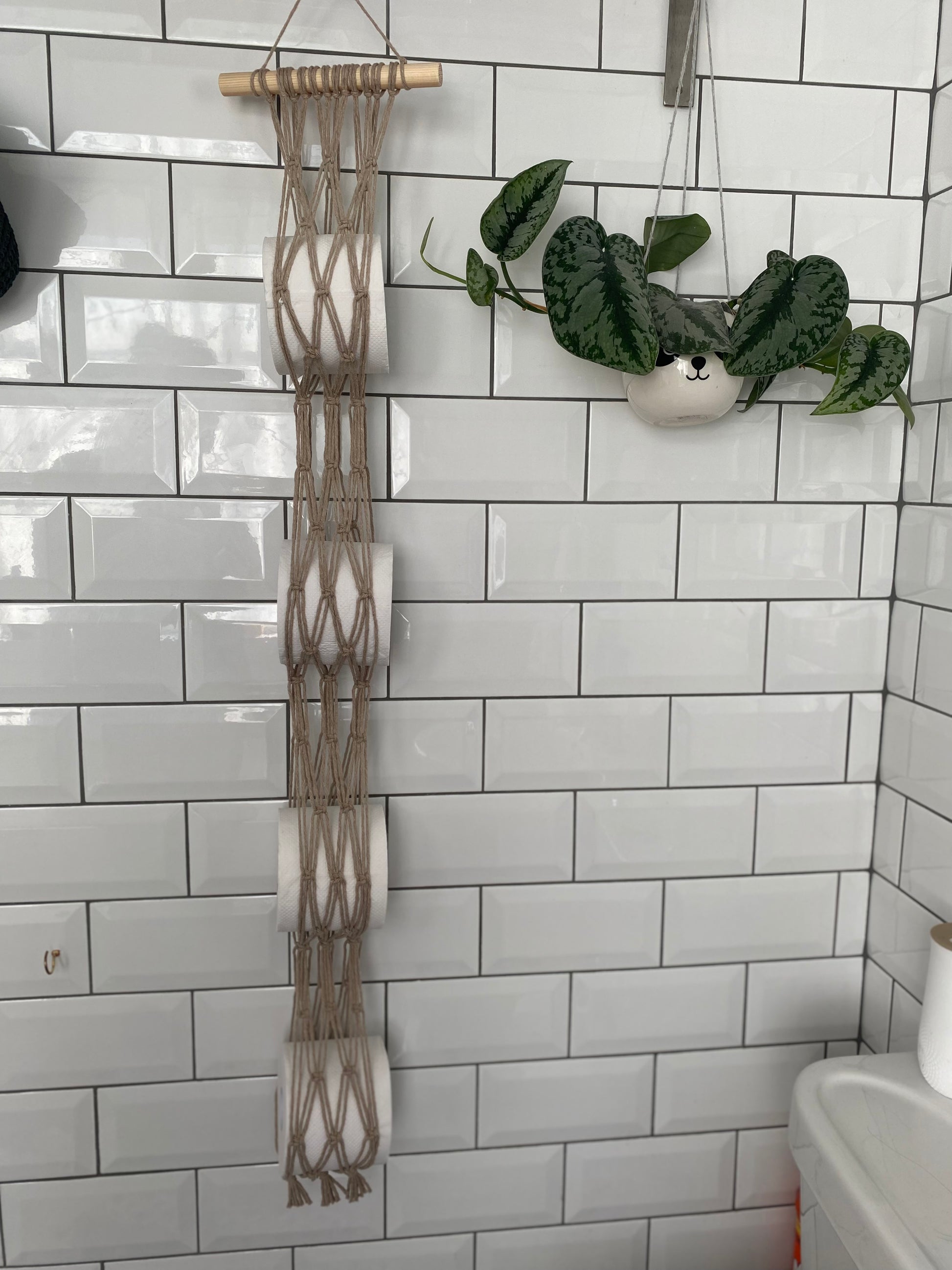 A white tiled wall features a Macra-Made-With-Love Toilet paper wall mounted storage crafted from recycled cotton, hanging on a wooden rod and holding four rolls. To the right, a small white hanging planter contains a leafy green plant. A white container is visible at the bottom right corner.