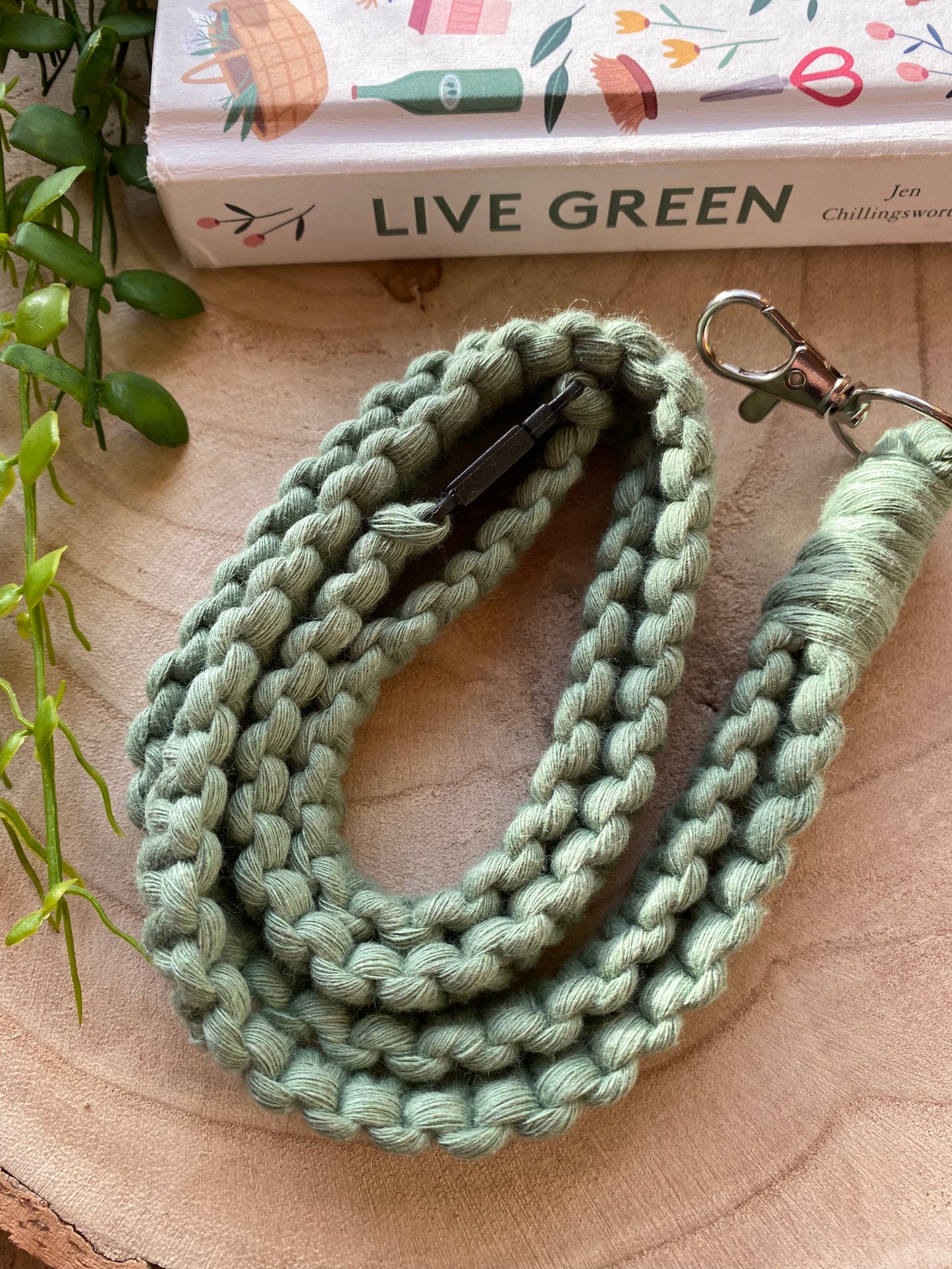 A green braided leash with a metal clip is coiled on a wooden surface. Beside it lies a colorful book titled "Live Green" by Jen Chillingsworth. Nearby, there is an Eco friendly lanyard badge holder - 34” from Macra-Made-With-Love and a small section of a trailing plant, enhancing the natural, eco-friendly theme of the scene.