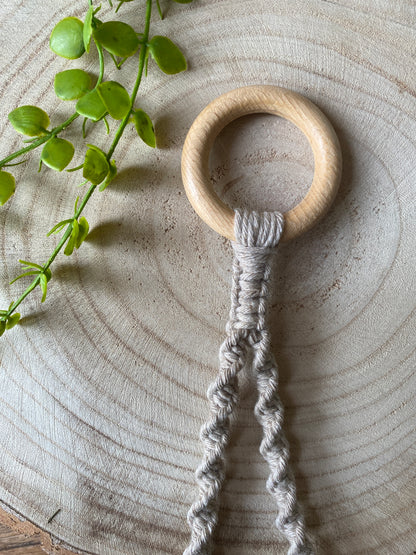 A Macra-Made-With-Love Single toilet roll holder with beige macramé knots hangs on a light wooden, circular surface. A green plant with small, oval leaves extends diagonally into the frame from the top left corner, providing a natural accent to this eco-friendly decor piece.