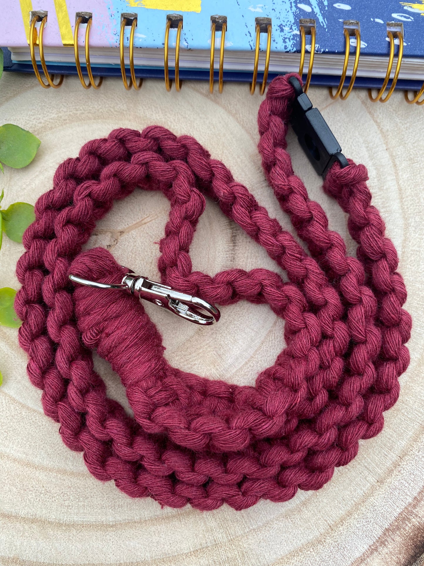 A handcrafted, braided maroon **Eco friendly lanyard badge holder - 34”** by **Macra-Made-With-Love** with a silver clasp is coiled neatly on a circular wooden surface. The background features part of a colorful, spiral-bound notebook with a few green leaves slightly visible on the left side.