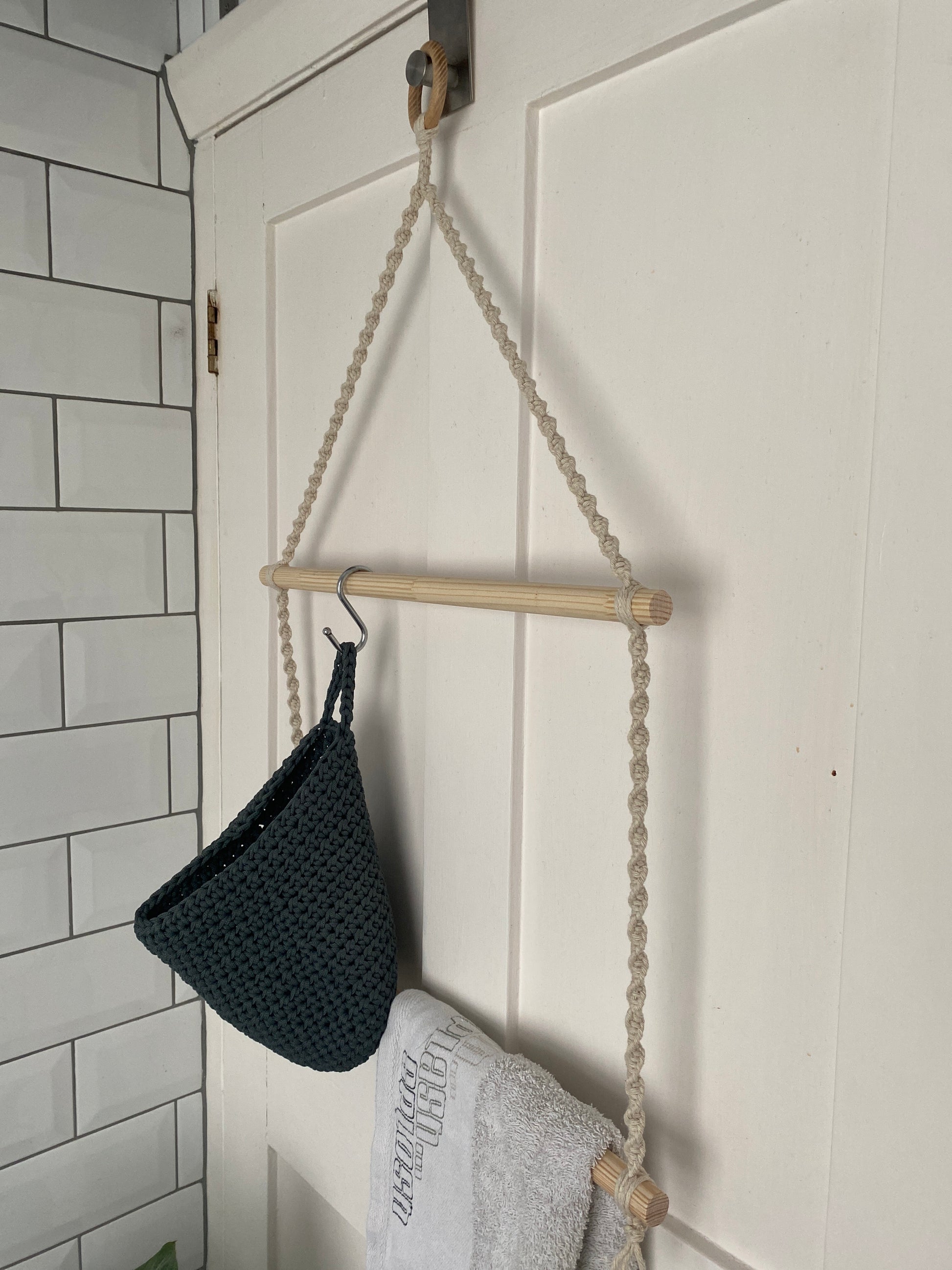 A white door features a handmade macrame towel holder by Macra-Made-With-Love, with a hanging wooden rod suspended by thick, braided rope. A dark, knitted basket hangs on a hook from the rod, and a gray towel with text embroidered on it is draped over the Two Tier Bathroom Towel Holder. White tiled wall with black grout lines seen to the left.