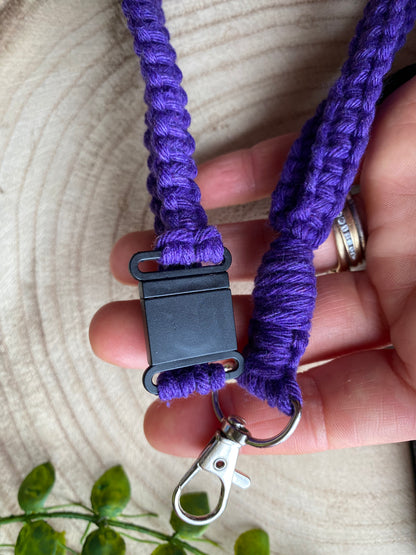 A close-up of a hand holding an Eco friendly lanyard badge holder - 34” by Macra-Made-With-Love, featuring a purple woven design with a black plastic breakaway clip and a metal lobster clasp, perfect as an ID badge holder. The background features a section of round wood and a few green leaves.