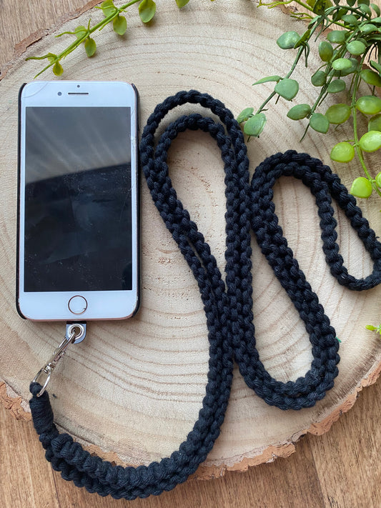 A smartphone with a white front and black screen is attached to a black, braided lanyard. This eco-friendly, handmade Recycled cotton crossbody phone strap by Macra-Made-With-Love is coiled in a loop around the phone. The items are placed on a round wooden surface with green leafy branches in the corners of the image.