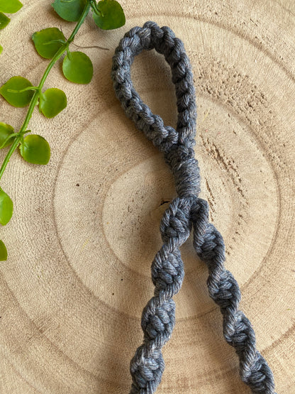 A handmade grey Single toilet roll holder from Macra-Made-With-Love with a loop at the top lies on a wooden surface. The holder, an eco-friendly decor piece, features a series of spiral knots. A small green plant with round leaves is partially visible on the left side of the image.