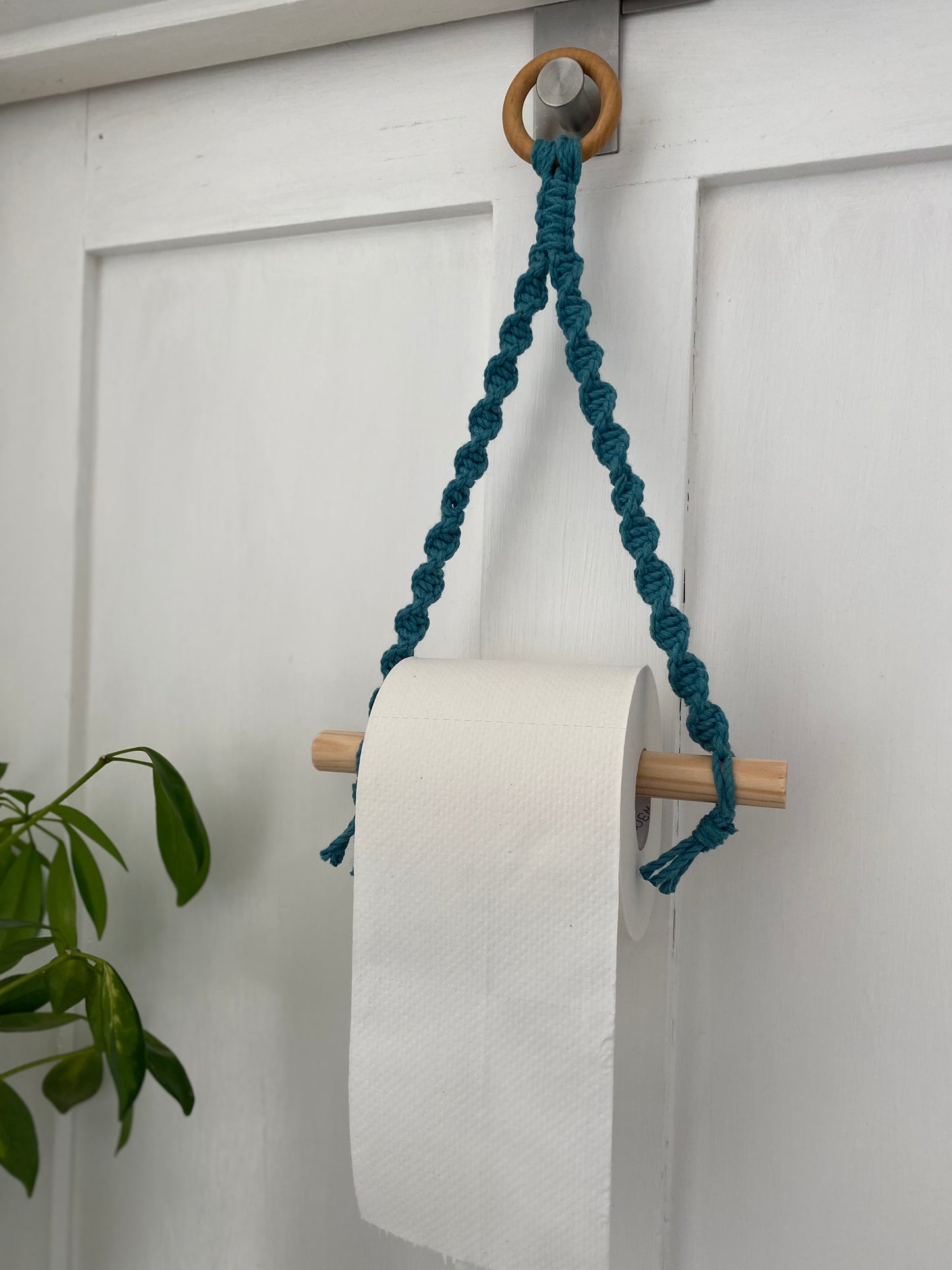 A wooden holder with a teal braided cord hangs from a metal hook on a white door. This Single toilet roll holder by Macra-Made-With-Love supports a roll of white toilet paper, and a green plant with broad leaves is partially visible to the left—an ideal bathroom accessory for eco-friendly decor.