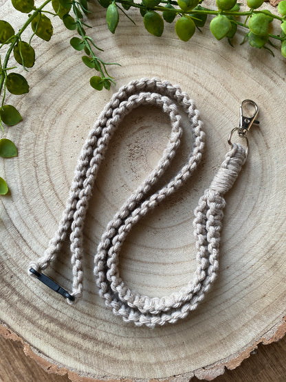 A handmade, braided beige macrame Eco friendly lanyard badge holder - 34” by Macra-Made-With-Love rests on a round wooden surface. Featuring a metal clip and loop, this eco-friendly lanyard is perfect as an ID badge holder. Greenery decorates the top left and top edge of the image.