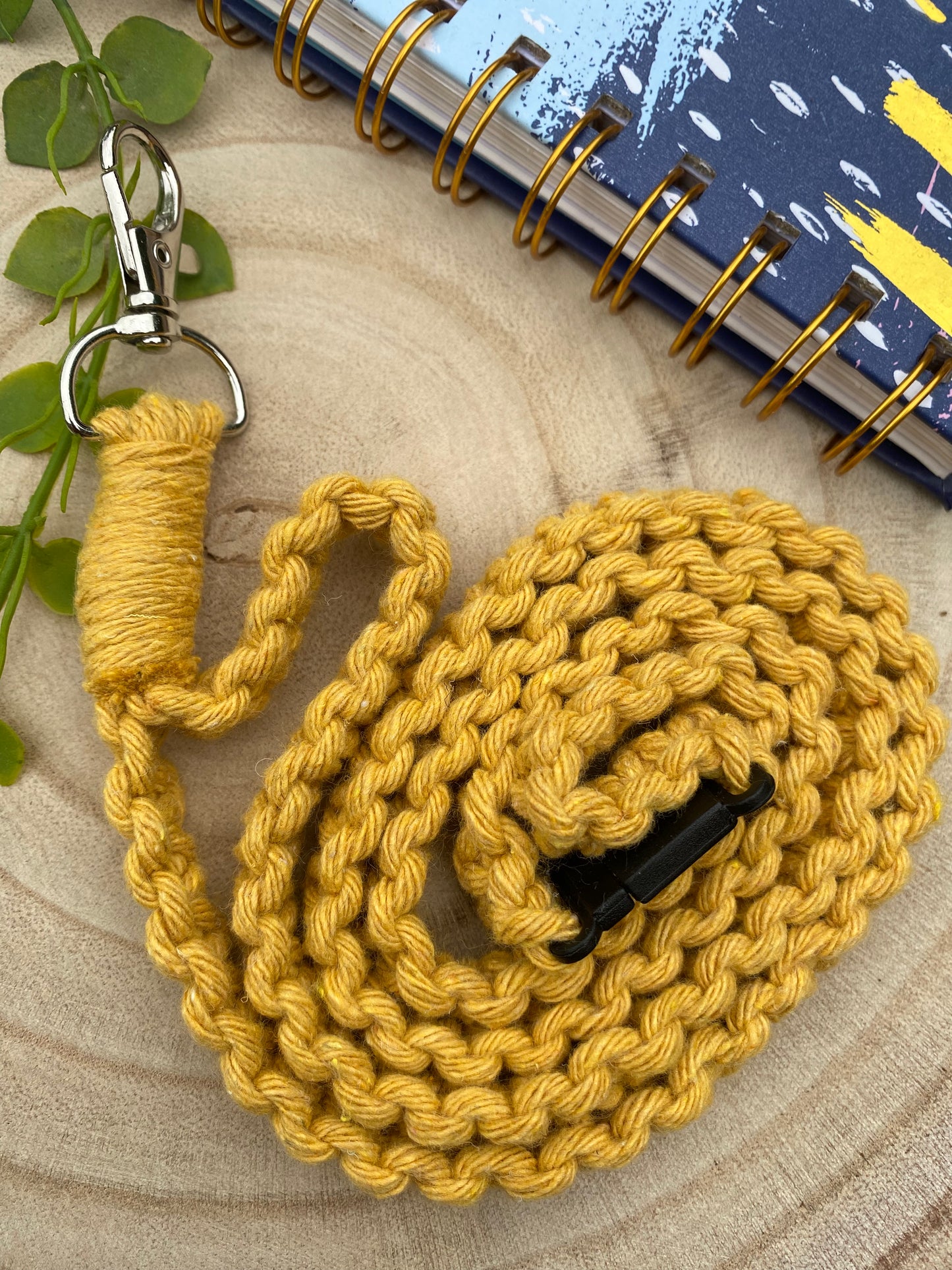 A yellow Eco friendly lanyard badge holder - 34” by Macra-Made-With-Love with a metal clip lies coiled on a light wooden surface. Part of a blue spiral-bound notebook with yellow and white patterns is visible in the top right corner. A small green plant leaf is placed to the left of the lanyard.