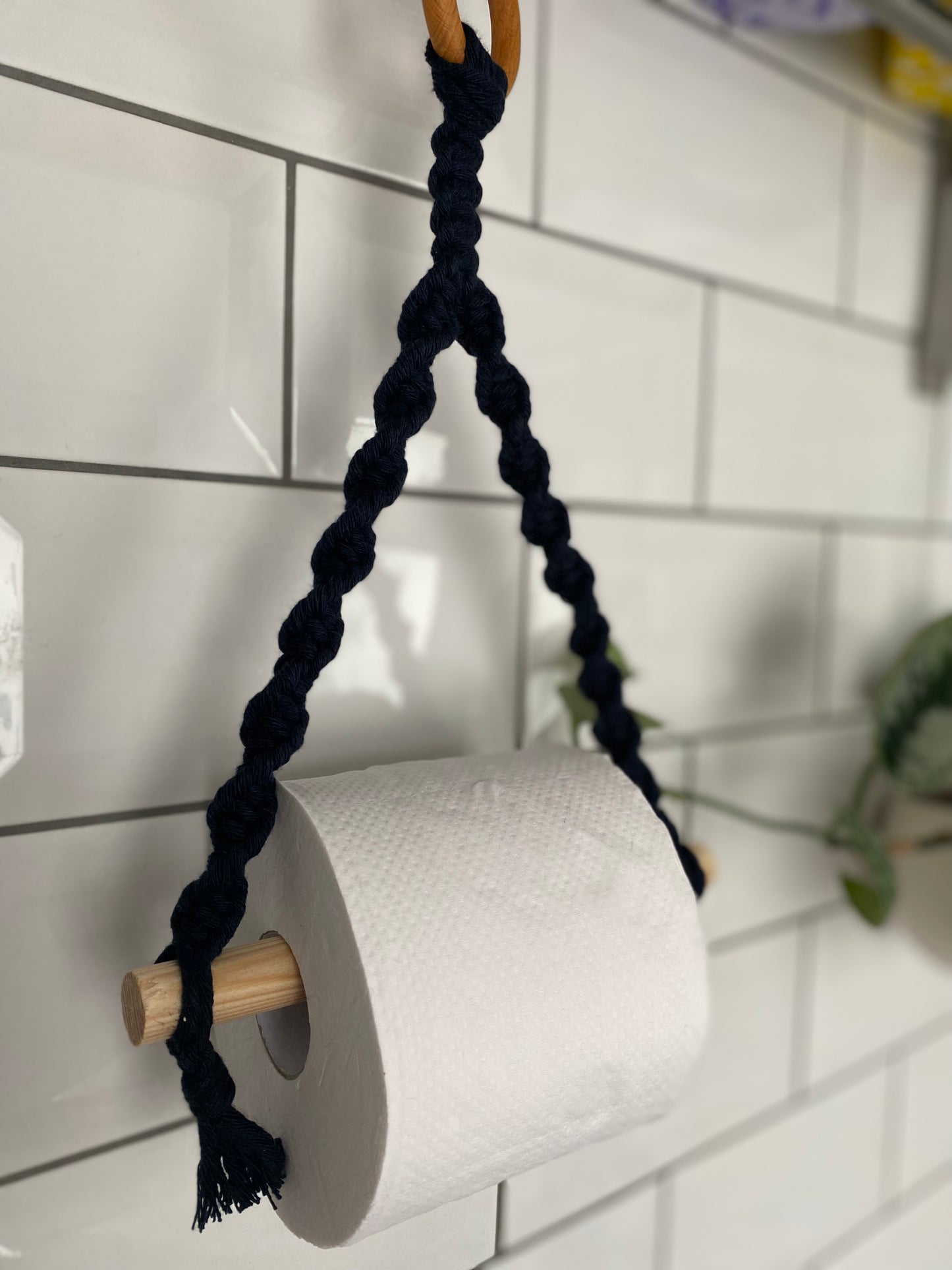 A roll of toilet paper hangs on a wooden dowel, showcasing a Macra-Made-With-Love Single toilet roll holder made from thick, twisted black rope attached to a wall tiled with white rectangular tiles. Some green plants are partially visible in the background, adding to the eco-friendly decor.