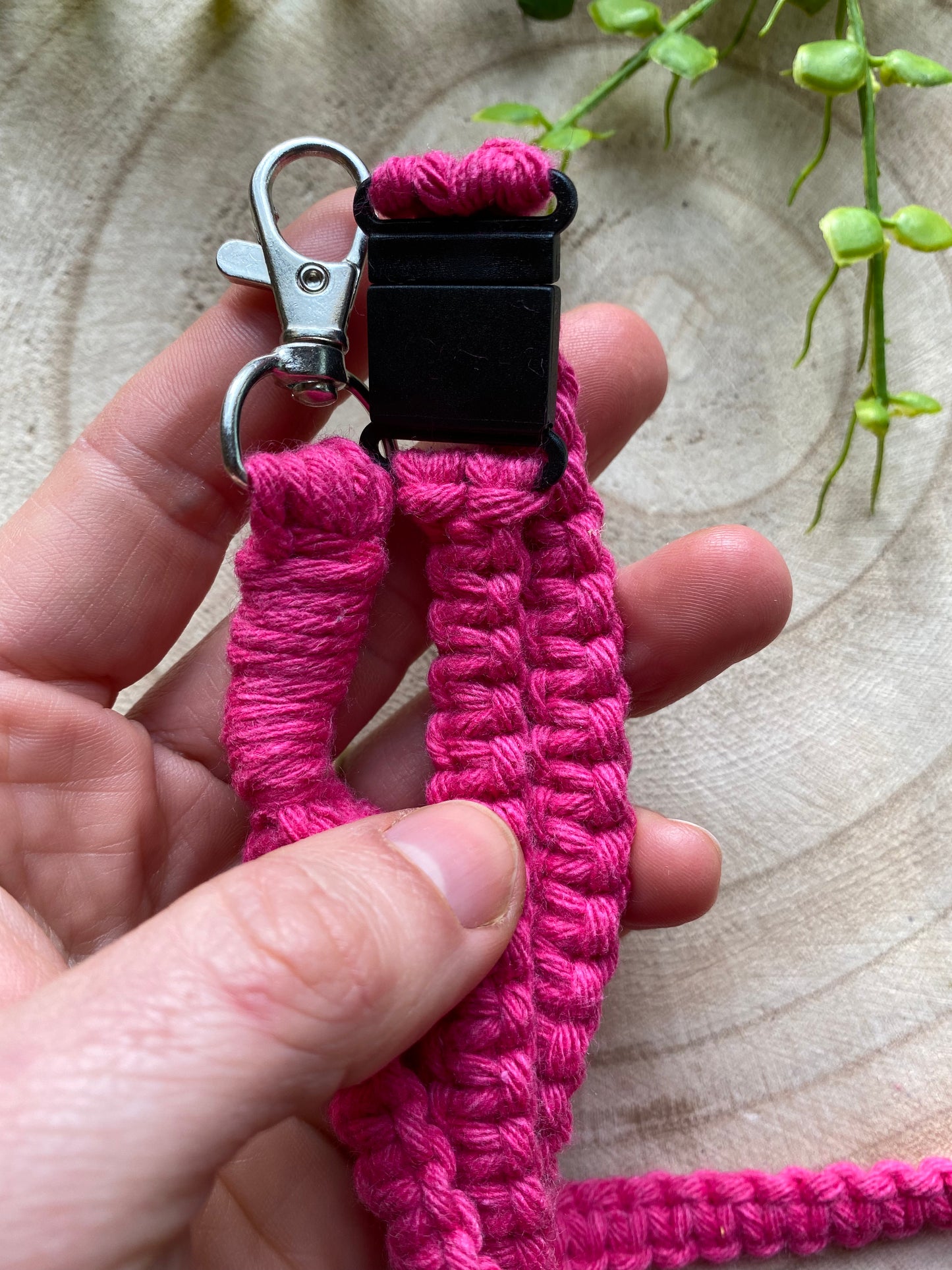 A hand holds a bright pink, woven, handmade lanyard with a metallic clip and black plastic buckle. The Eco friendly lanyard badge holder - 34” from Macra-Made-With-Love is set against a circular wooden surface adorned with some green vine-like plants.