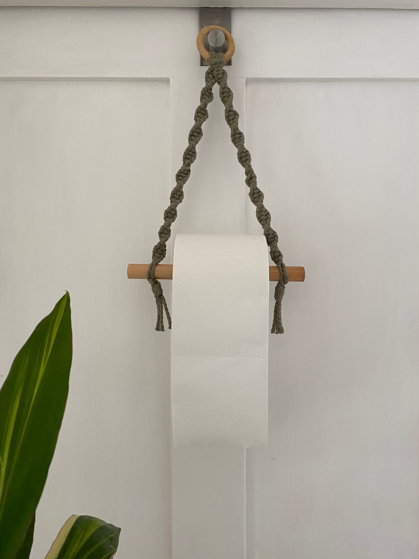 A Single toilet roll holder by Macra-Made-With-Love is hung on a wooden dowel, which is suspended by a handmade macrame toilet roll holder mounted on a white door. A green plant's leaves are visible in the lower left corner of the image, completing the eco-friendly decor.