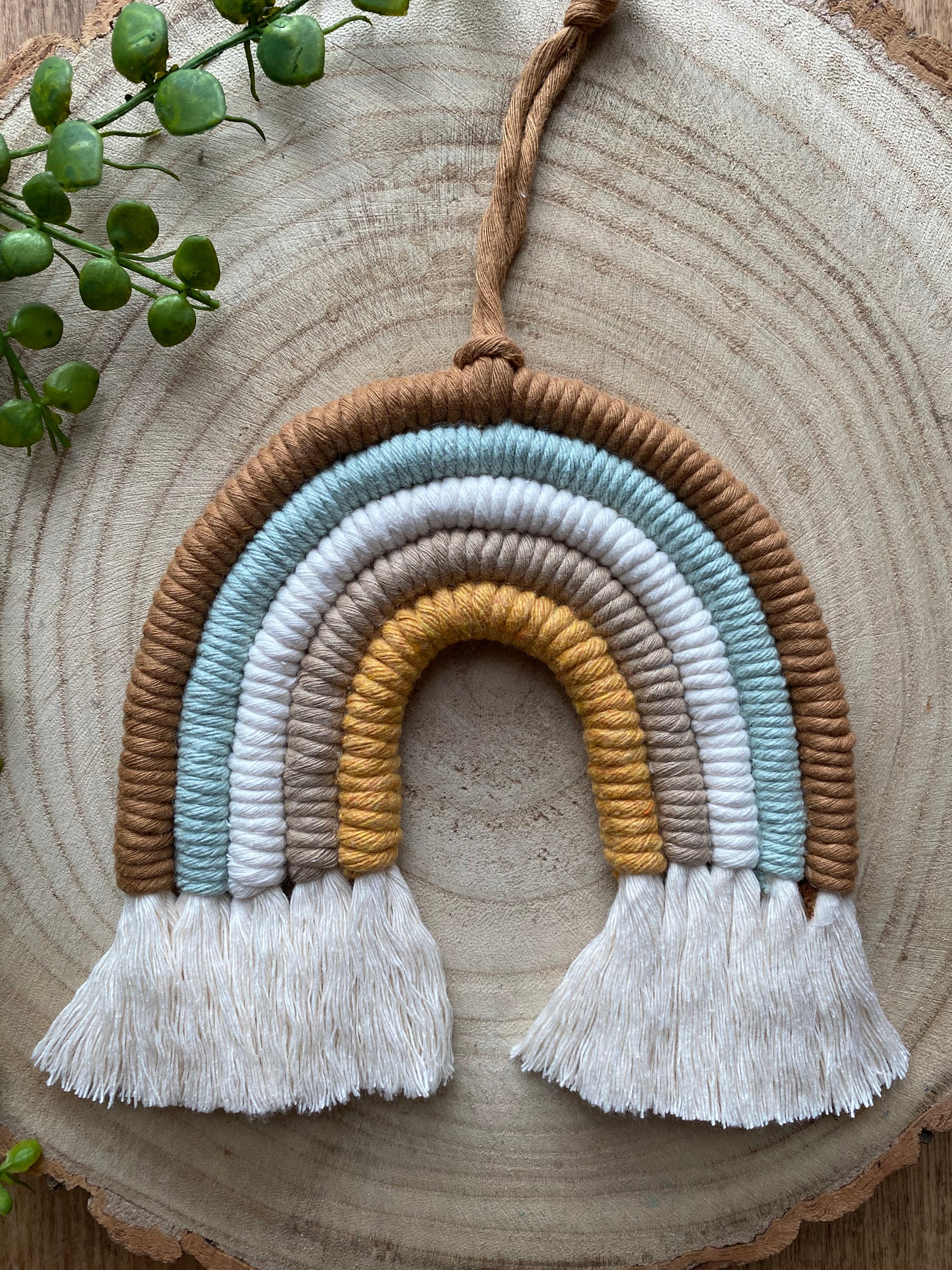 A handmade Macra-Made-With-Love neutral rainbow with five colored bands (brown, light blue, white, ochre, and gray) hanging on a wooden background. The neutral rainbow has white fringes at both ends. A small green plant is seen in the top left corner. Perfect as a baby shower gift made with sustainable practices.