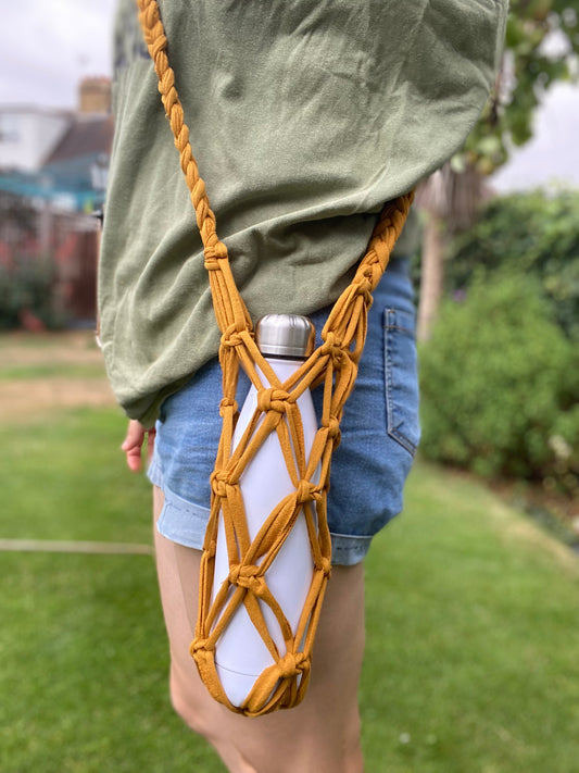 A person wearing an olive-green shirt and denim shorts carries a white water bottle in an Eco friendly crossbody reusable bottle holder by Macra-Made-With-Love in a brown macrame sling over their shoulder. The scene is set outdoors with a green grassy background and some blurred elements like trees and buildings.