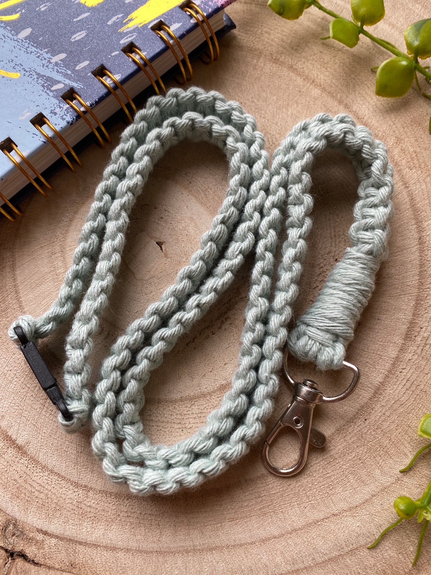 A light-colored, handmade Eco friendly lanyard badge holder - 34” by Macra-Made-With-Love with a metal clip is coiled on a wooden surface. A notebook with a spiral binding and colorful abstract cover, along with a vine plant, are partially visible in the background.