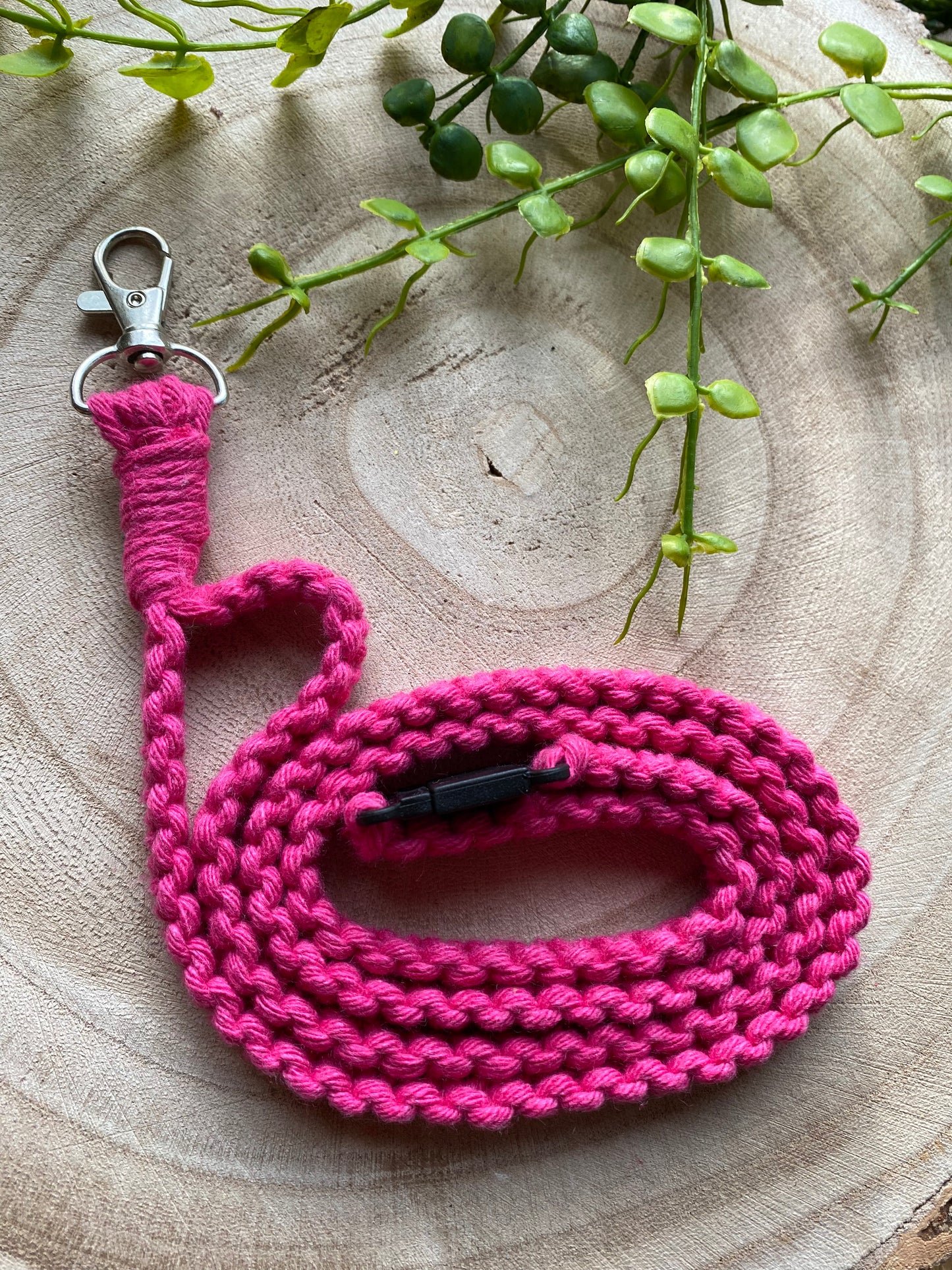 A vibrant pink Eco friendly lanyard badge holder - 34” from Macra-Made-With-Love with a metal clasp is coiled neatly on a circular wooden surface. A few green leafy plants are placed in the background, adding a natural touch to the composition. This handmade lanyard-inspired leash combines style with eco-friendly materials for your furry friend.