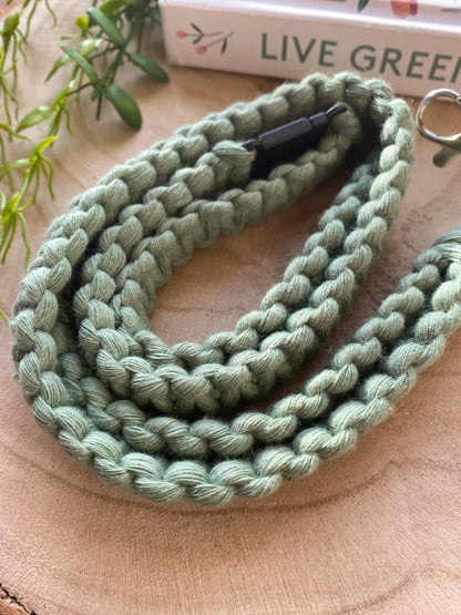 Close-up of a green, braided fabric **Eco friendly lanyard badge holder - 34”** from **Macra-Made-With-Love** coiled on a wooden surface. The lanyard appears sturdy, with a metal clasp partially visible. In the background, part of a book with "LIVE GREEN" on the spine and some green plant leaves can be seen. This eco-friendly lanyard adds a touch of nature to your gear.